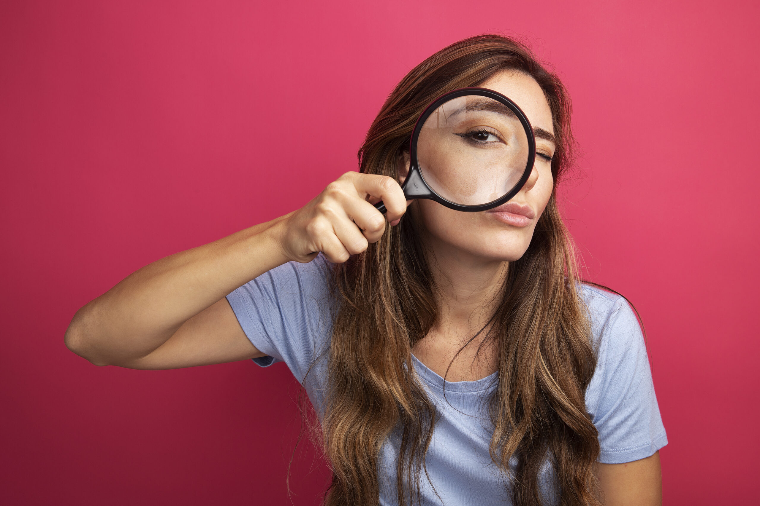 young beautiful woman in blue t-shirt looking at camera through magnifying glass with interest standing over pink background
